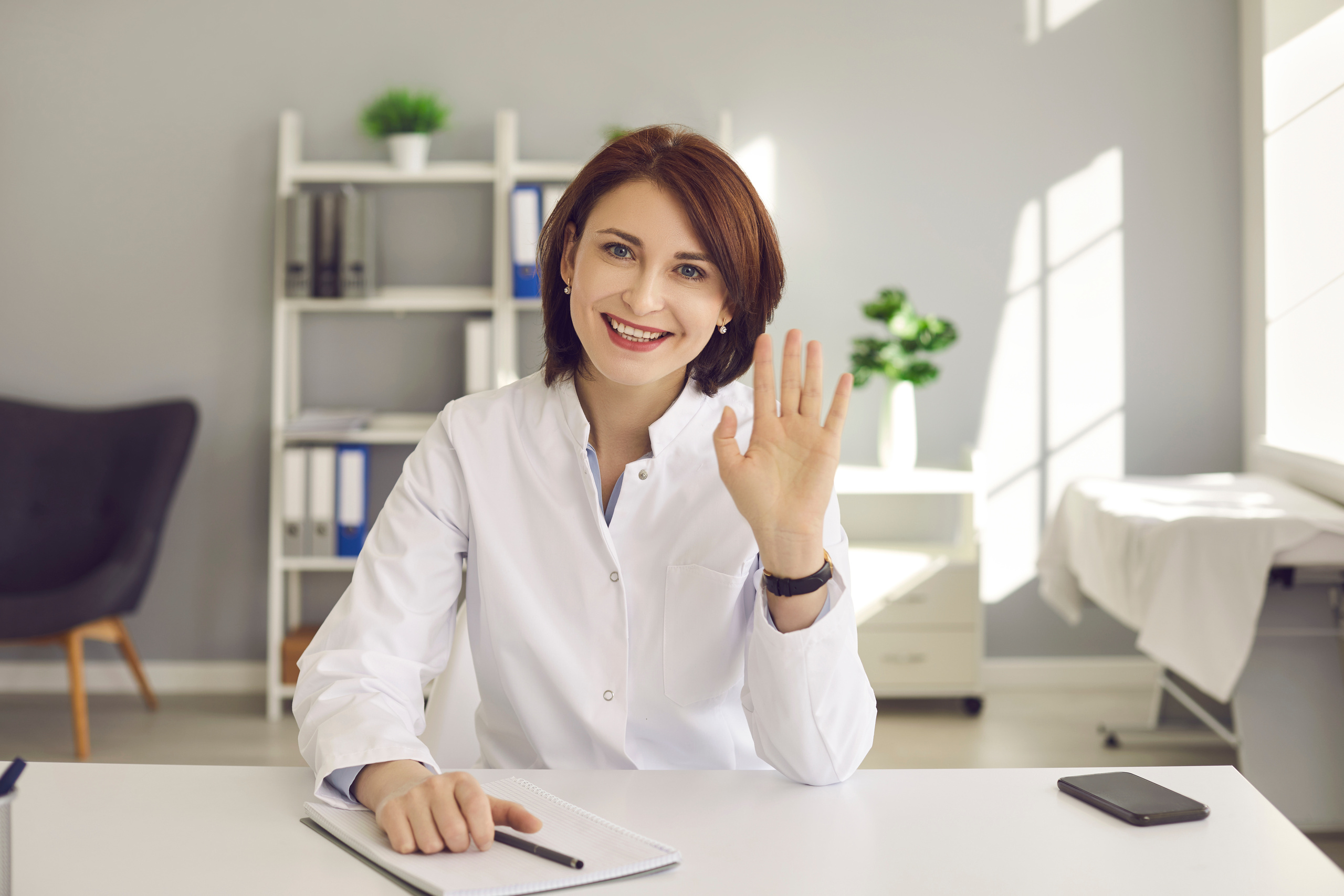 Friendly Middle-Aged Female Doctor Waving Hand Greeting a Patient via Video Link.